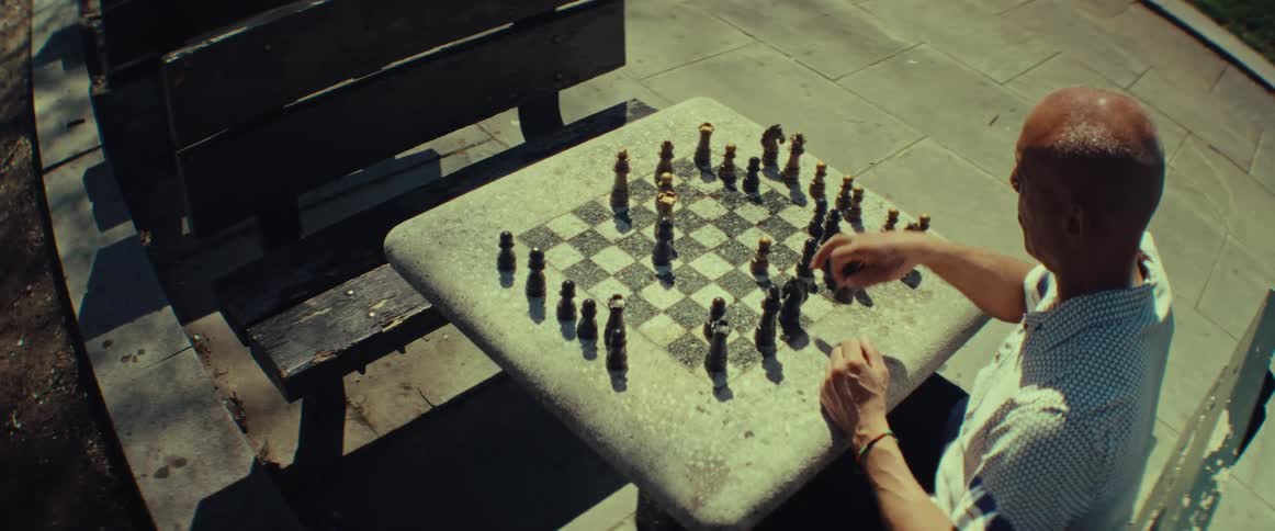 An older man plays a thoughtful game of chess at a stone table in a sunlit park