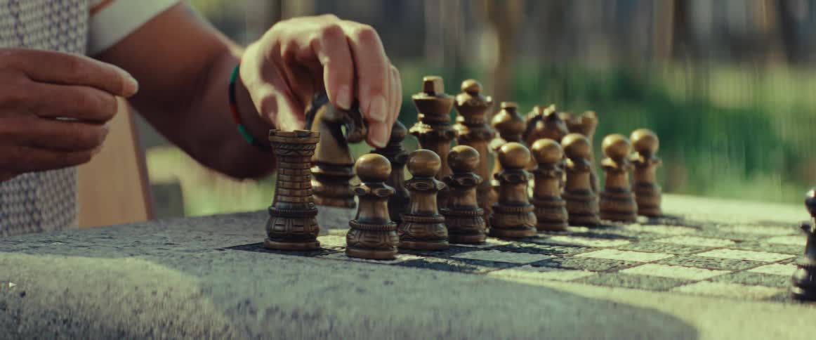A close-up of a person's hands thoughtfully setting up wooden chess pieces on a stone chessboard outdoors