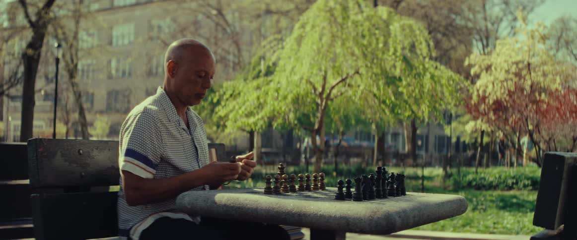 An elderly man patiently sets up a chessboard on a stone table in a peaceful city park