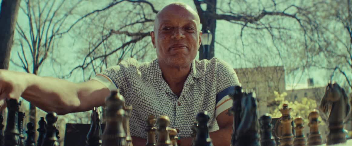 A smiling man makes a confident move while playing a game of chess in a park, shot from the opponent's perspective