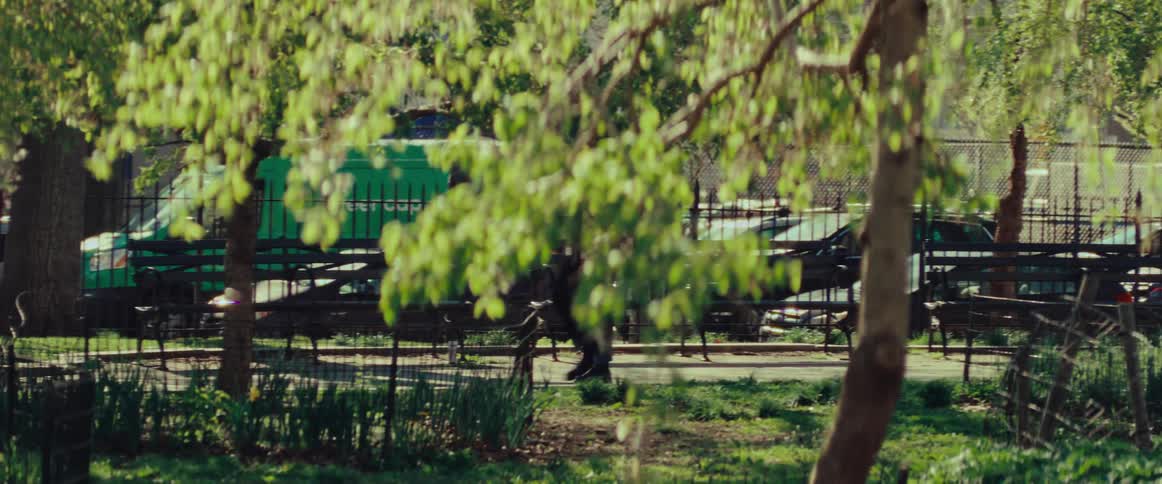 An older man carries a chessboard while strolling through a sunlit city park on a bright spring day