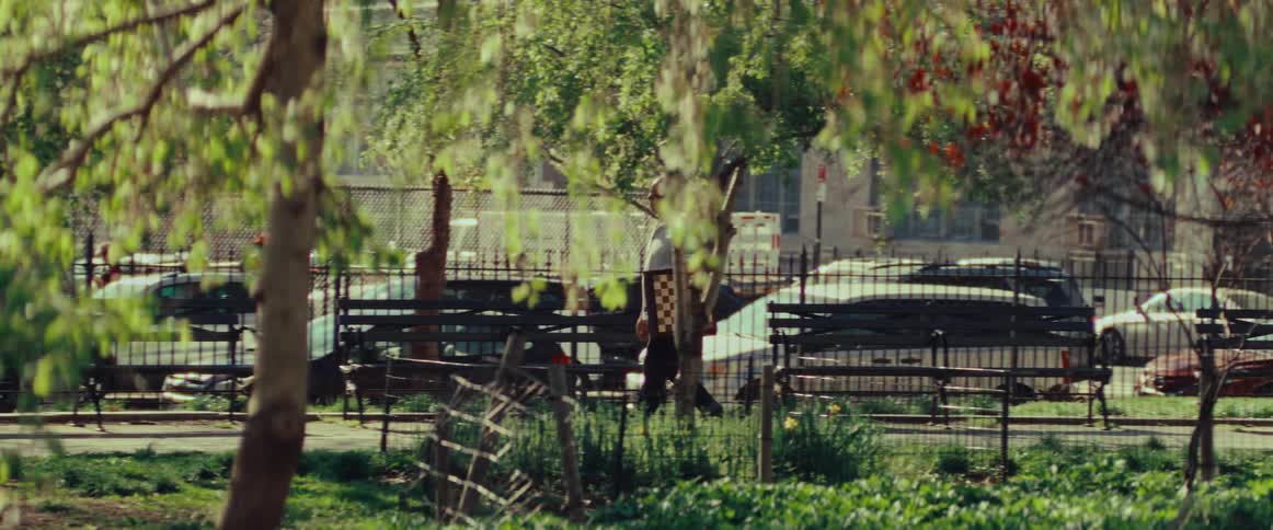 Slow motion shot of a man carrying a chessboard as he walks through a sunny urban park with lush green trees