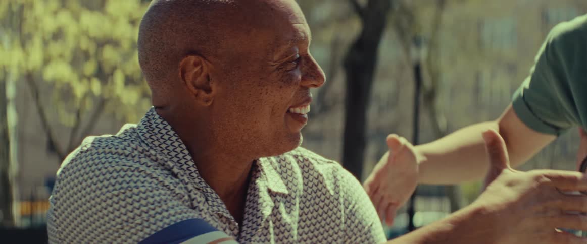 A smiling older man enjoys a conversation and shakes hands with a friend in a sunlit city park