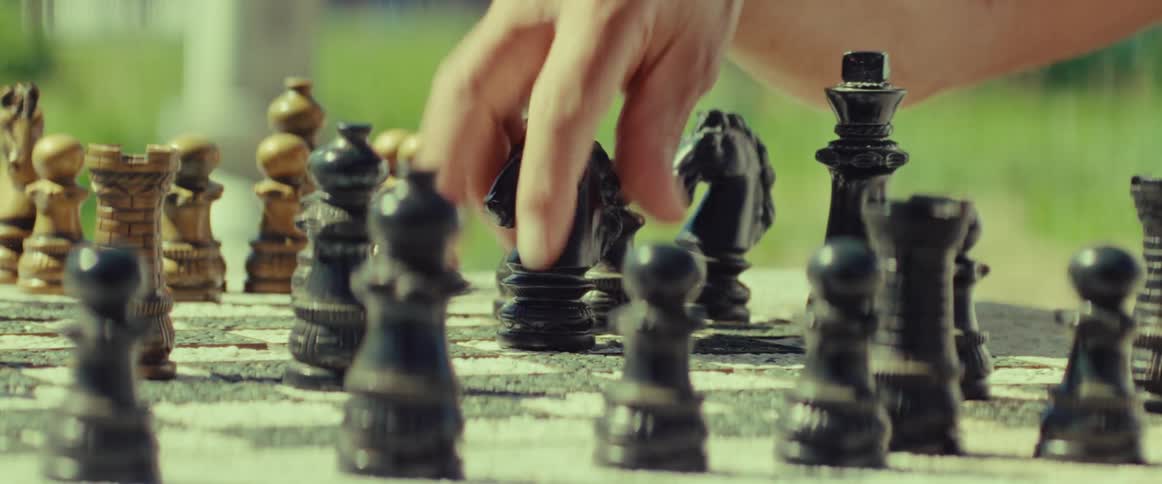 A close-up shot of a player moving a black knight piece on an ornate chessboard outdoors