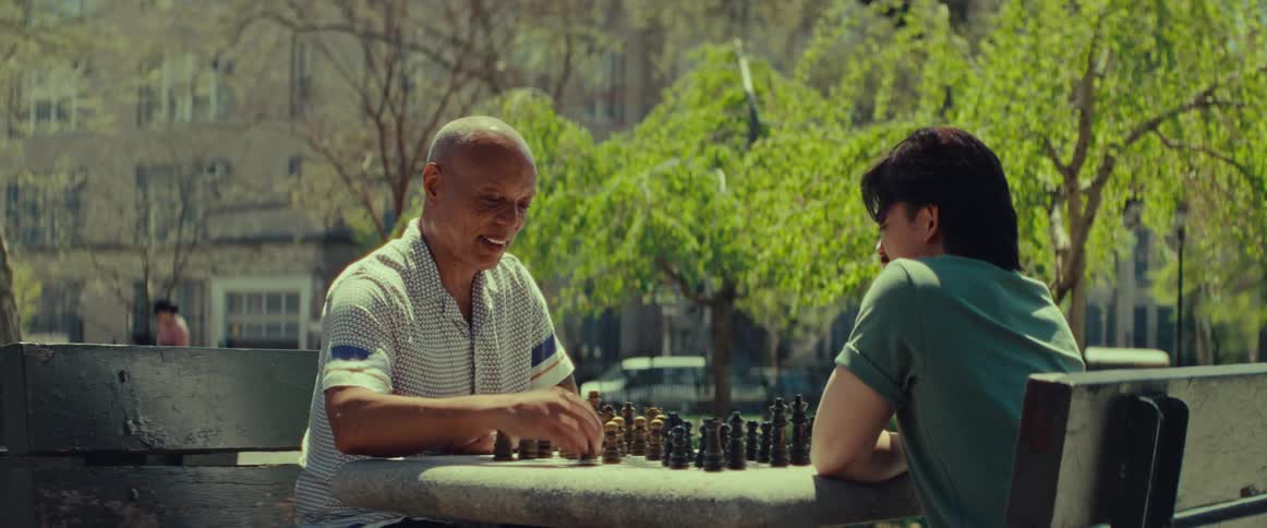 An older man smiles and makes a move while playing a friendly game of chess with a young man in a sunlit park