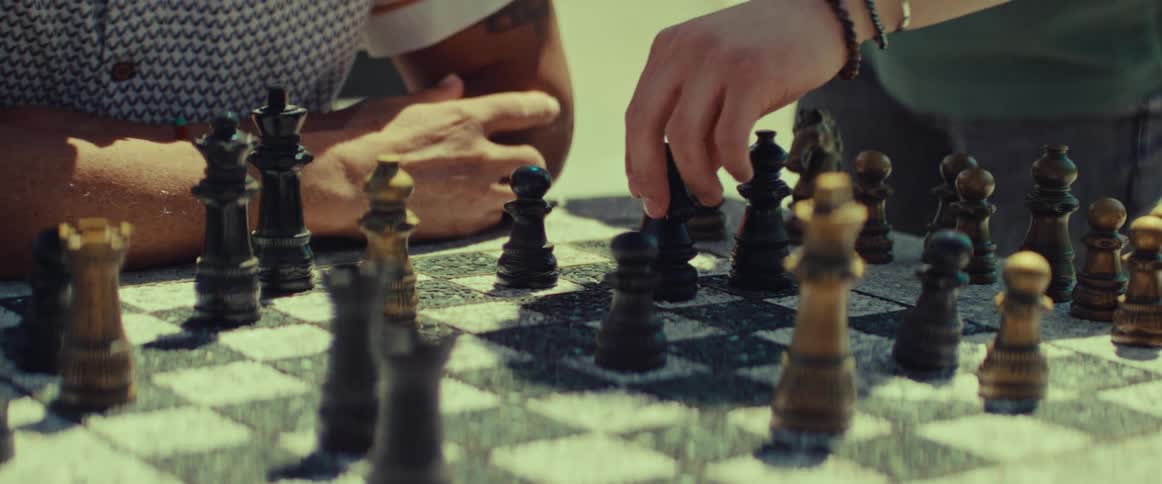 A cinematic close-up of a hand moving a knight during a game of chess played outdoors on a sunny day