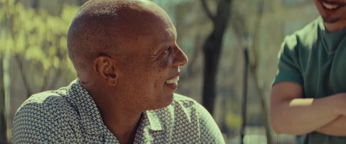 An older Black man smiles warmly and shakes hands with a friend while enjoying a sunny day in the park