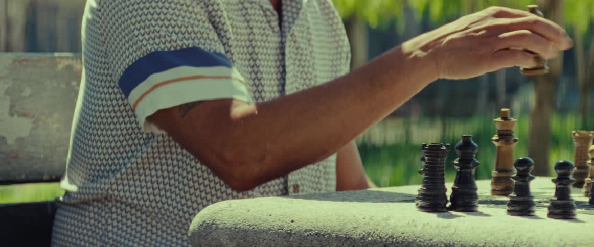 A panning shot reveals a man making a move during a game of chess at a stone table in a sunny park