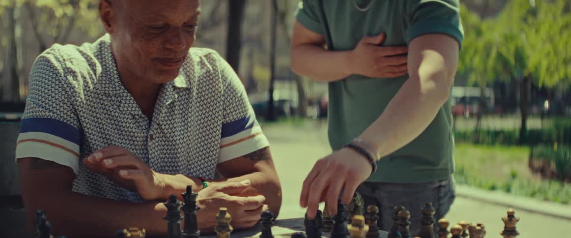 Two men share a friendly laugh and handshake after finishing a game of chess in a sunny city park
