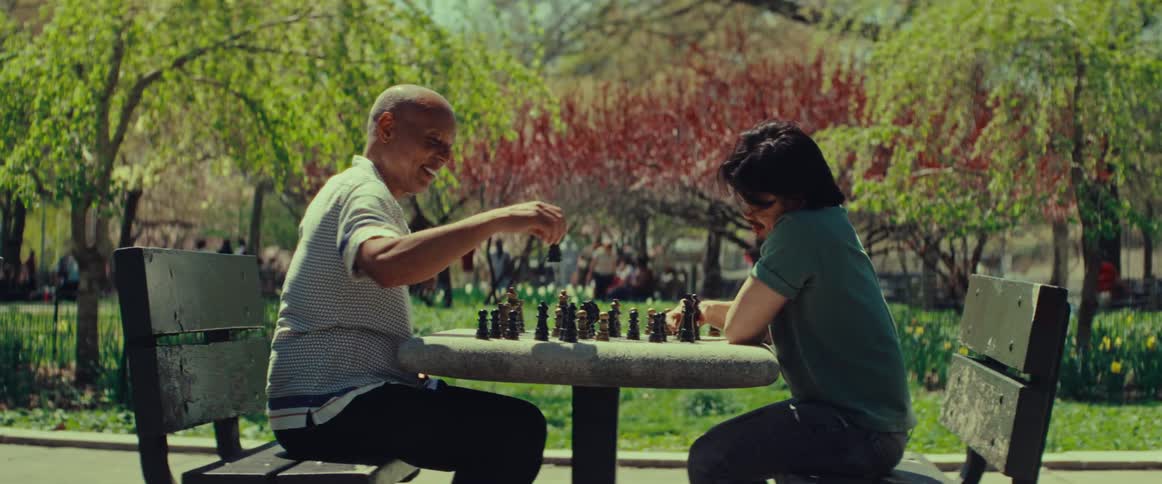 An older man smiles as he plays a friendly game of chess with a young man in a sunny city park