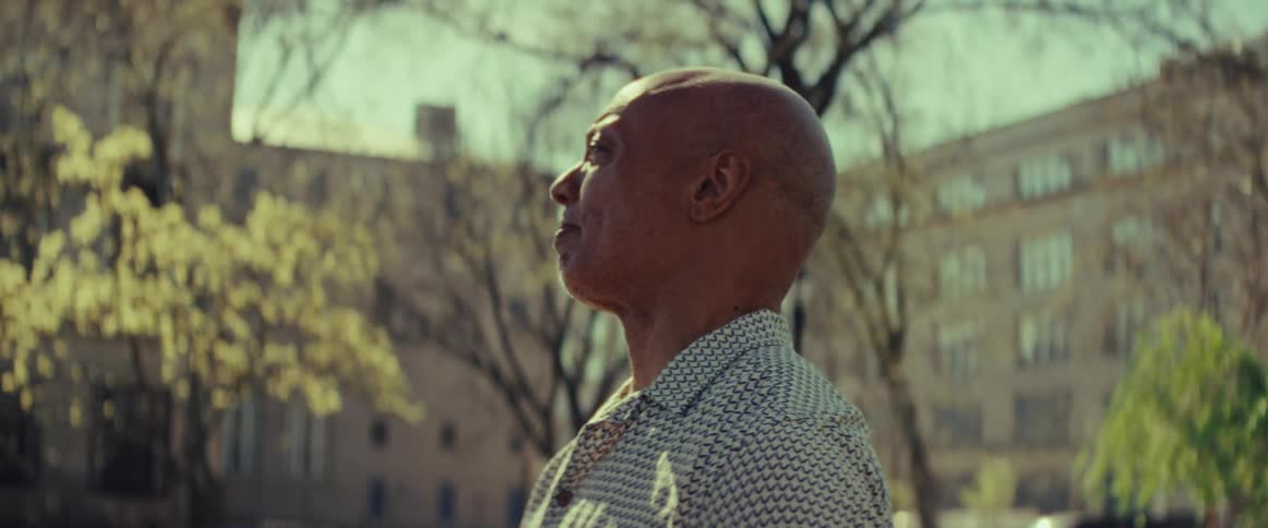 A contemplative elderly man holds a chessboard while soaking up the sun in a city park