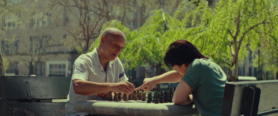 Two men play a friendly game of chess at a stone table in a sunny city park with lush green trees