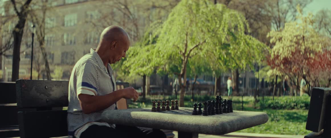A man sits alone at a chess table in a sunny city park, thoughtfully setting up the game pieces