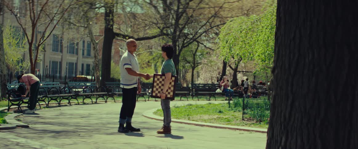 A young man holding a chessboard shakes hands with an older man in a sunny city park before they both walk away