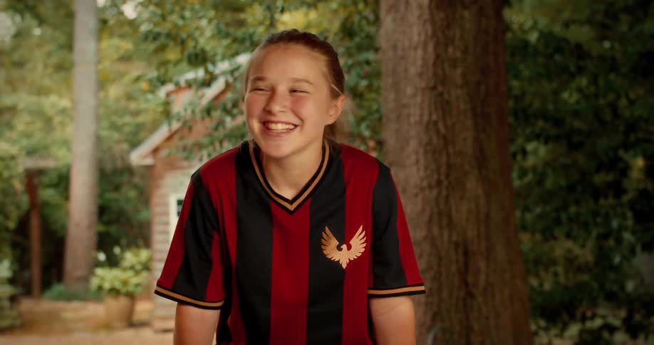 A young girl in a soccer uniform bursts into a fit of hysterical laughter while standing outdoors