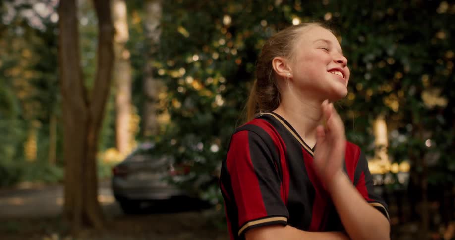 A young girl in a soccer uniform laughs heartily before becoming serious and thoughtful while standing outside