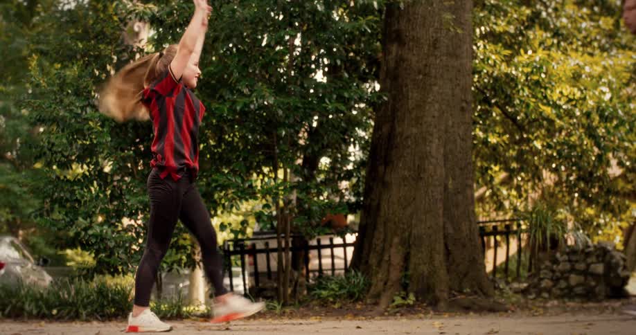 A young girl in a red and black striped shirt performs a slow-motion back walkover and cartwheel in a park as a man in a red tracksuit watches