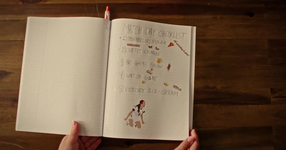 A young female soccer fan opens her decorated notebook to reveal a handwritten 'Match Day Checklist' on a wooden table