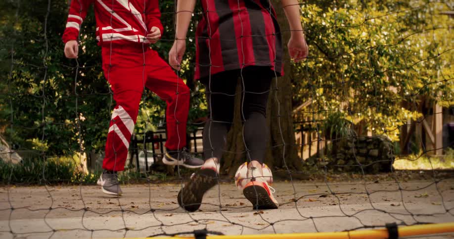 A father coaches his daughter in a one-on-one soccer drill in their driveway, captured in slow motion from behind the net