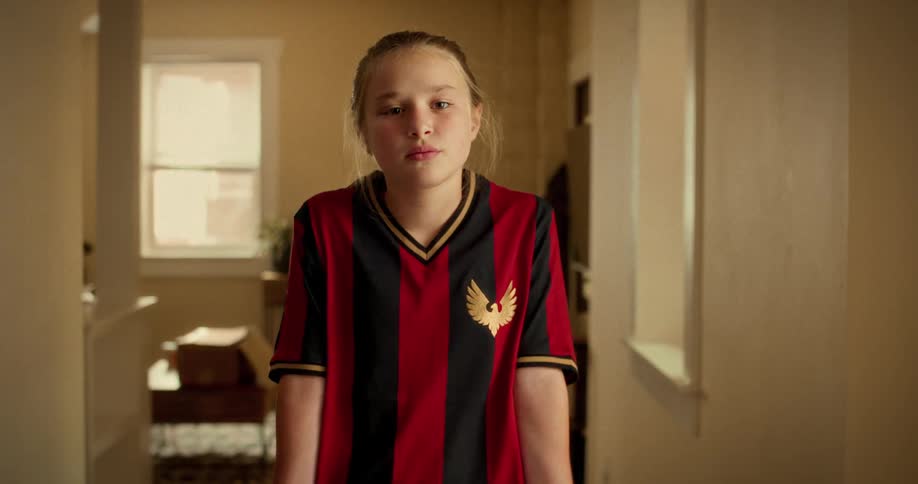 A sad young girl in a soccer uniform looks up with a determined expression while standing in a hallway