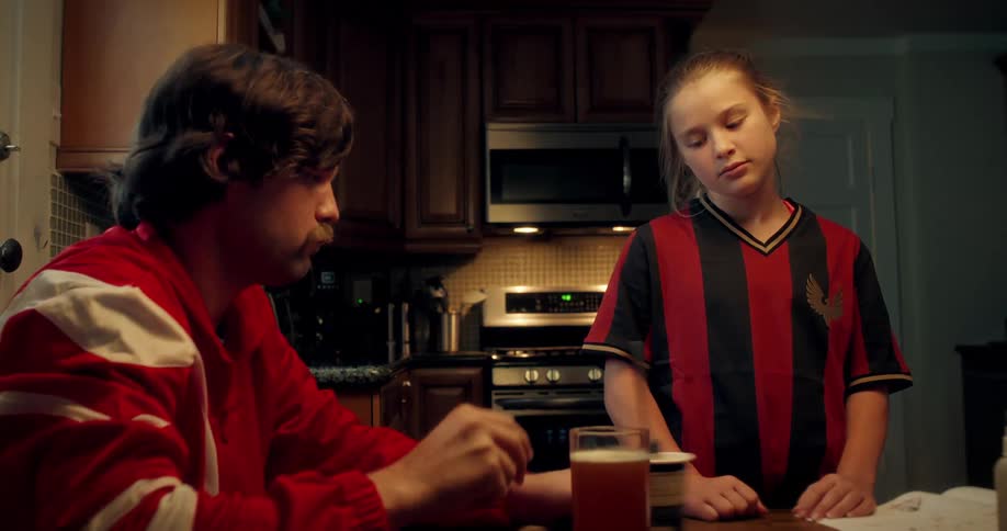A father in a red tracksuit sits at the kitchen table and talks to his sad daughter who is wearing a soccer jersey