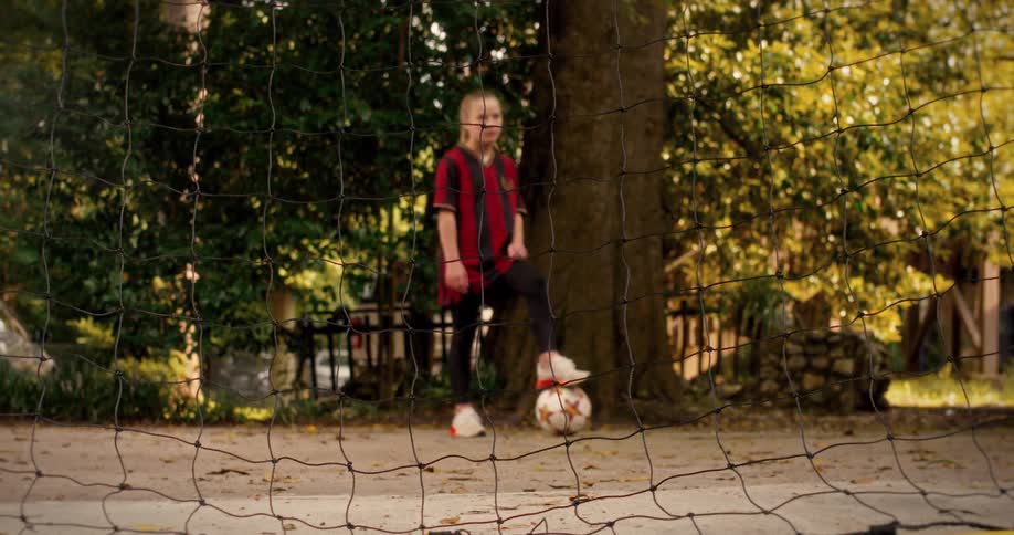 View through the soccer net of a determined young girl in her uniform, ready to play with her foot on the ball