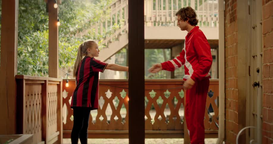 A young girl in a soccer uniform asks her father in a red tracksuit to take her picture with a smartphone on their back porch