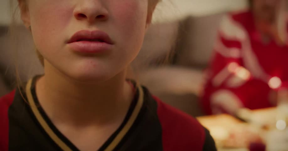 Close-up of a serious young girl in a sports jersey sitting indoors with a blurred family meal in the background