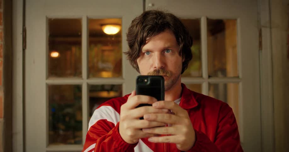 A mustached man in a retro tracksuit jacket intently scrolls through his smartphone indoors