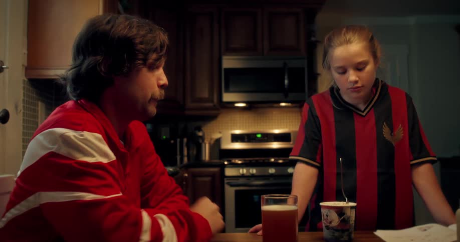 A father comforts his sad daughter in her soccer jersey as they talk in the kitchen at night