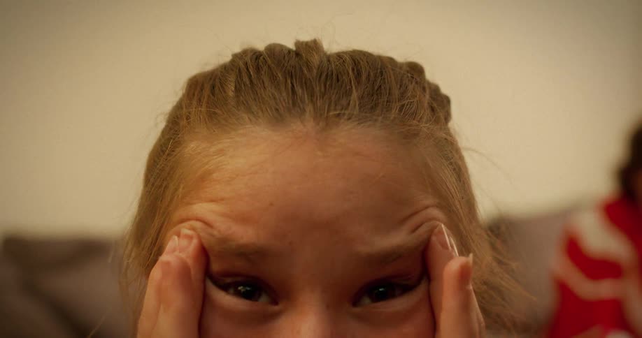 A young girl with braided hair playfully pulls at her eyes and wrinkles her forehead in a silly expression