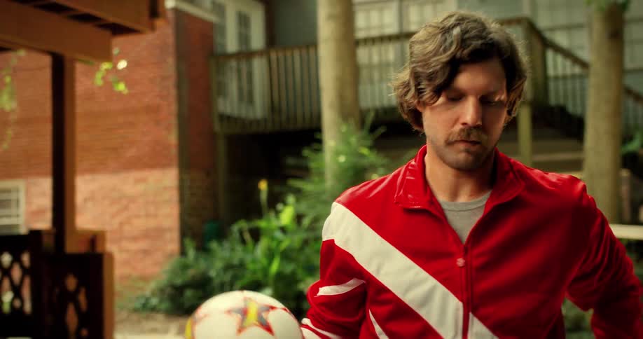 A man with a mustache in a vintage red tracksuit spins a soccer ball in slow motion before looking off thoughtfully