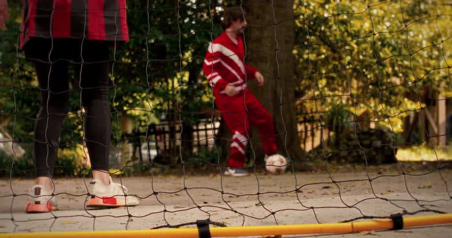 Through the net, a man in a red tracksuit juggles a soccer ball in slow motion during a casual game in the park