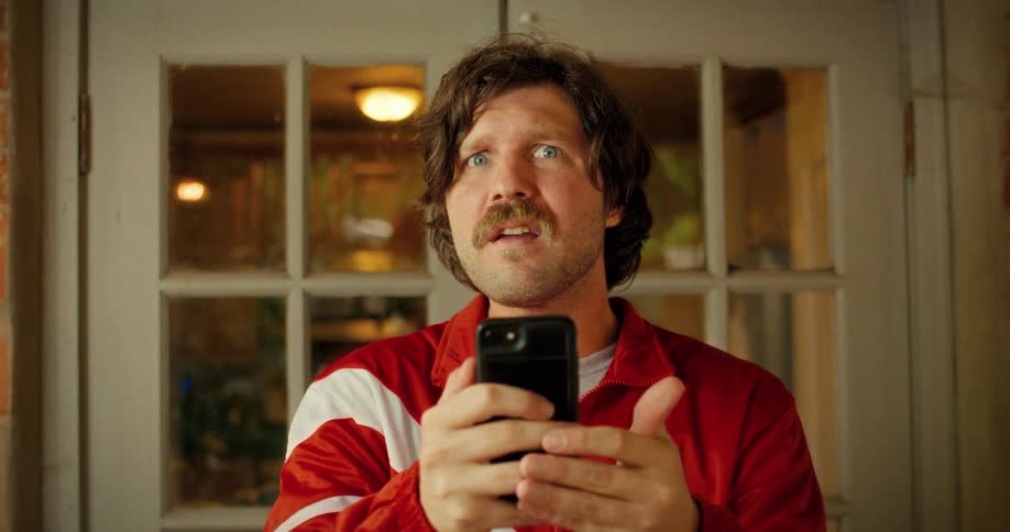 Close up of a concerned man in an 80s style tracksuit reading a shocking message on his smartphone