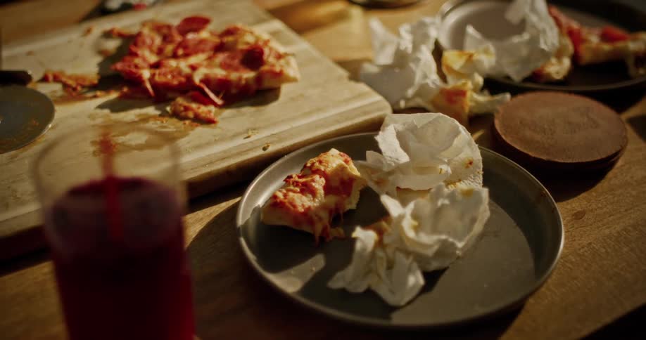 A messy wooden table shows the remnants of a pepperoni pizza dinner with dirty plates and crumpled napkins