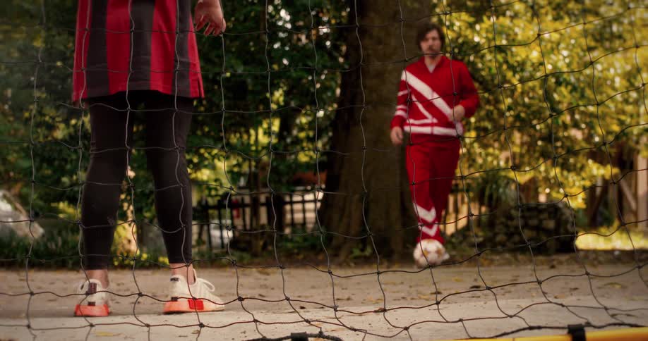 Slow motion shot from behind the goal as a man in a red tracksuit dribbles and kicks a soccer ball, scoring against his opponent