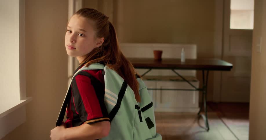 A young girl with a backpack looks over her shoulder with a serious expression before leaving home