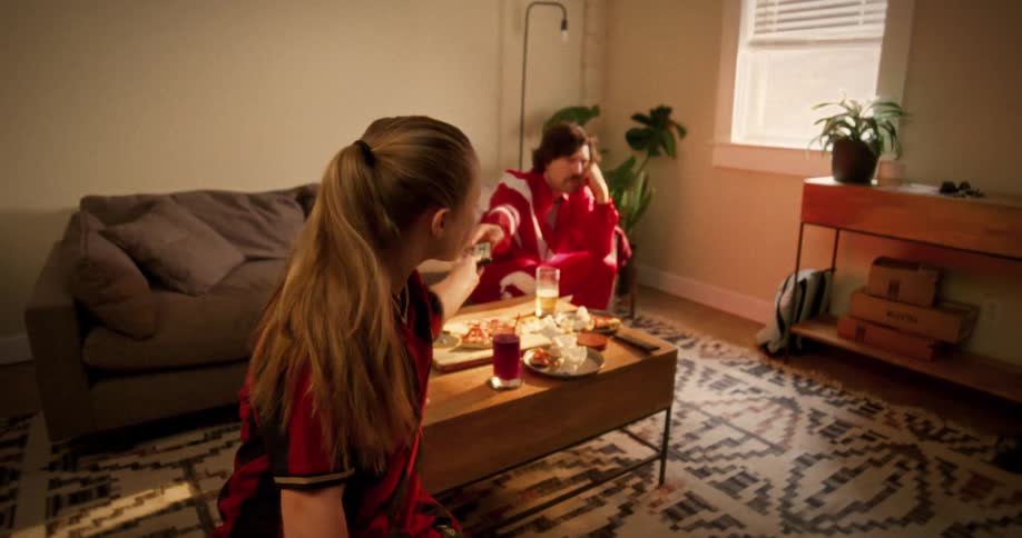 A young girl in a soccer jersey gets the TV remote from her disinterested father, puts in the batteries, and starts watching TV in the living room
