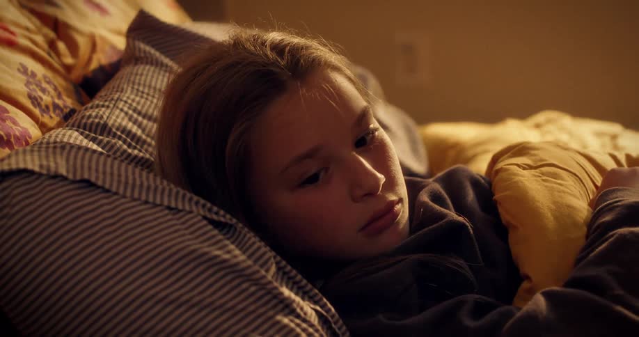 A close-up of a pensive young girl lying in bed, her face illuminated by warm, soft light as she contemplates