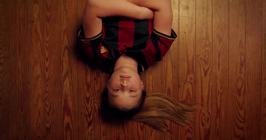 A thoughtful young girl in a soccer uniform lies on her back on a hardwood floor, looking up at the camera from a top-down perspective