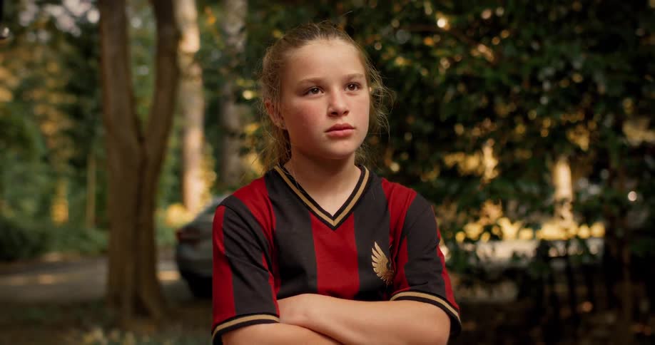 A serious young female soccer player stands with her arms crossed, looking off-camera with a determined expression
