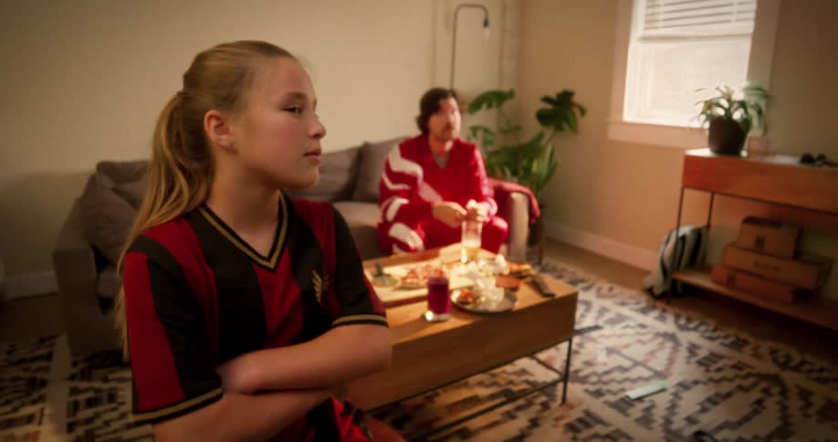 A serious young girl in a soccer jersey throws the remote and looks directly at the camera while watching a game with her dad