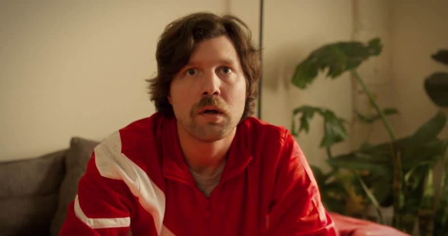 A mustached man in a retro red tracksuit goes from bored to shocked while watching something off-screen