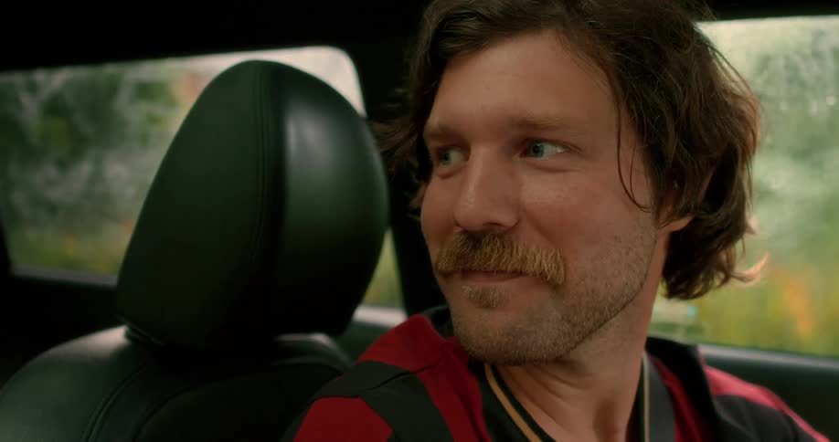 A smiling man with a mustache looks over at the driver from the passenger seat of a car