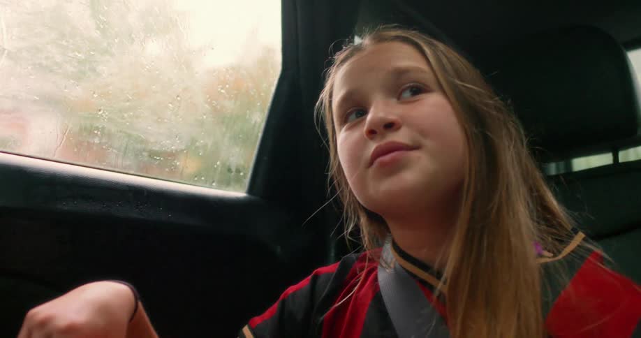 A thoughtful young girl in a sports jersey daydreams while riding in the back seat of a car on a rainy day