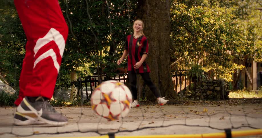A young girl in a soccer uniform happily kicks a soccer ball towards her coach during a sunny outdoor practice session in slow motion