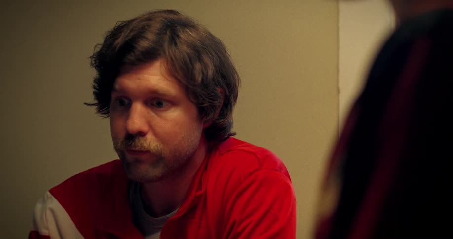 A melancholy man with a mustache, dressed in a retro red track jacket, slowly drinks a glass of beer