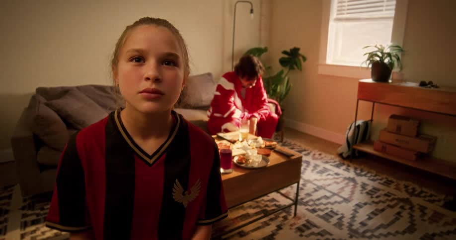 A young girl in a soccer jersey watches an intense game on TV with her father sitting on the couch in the background
