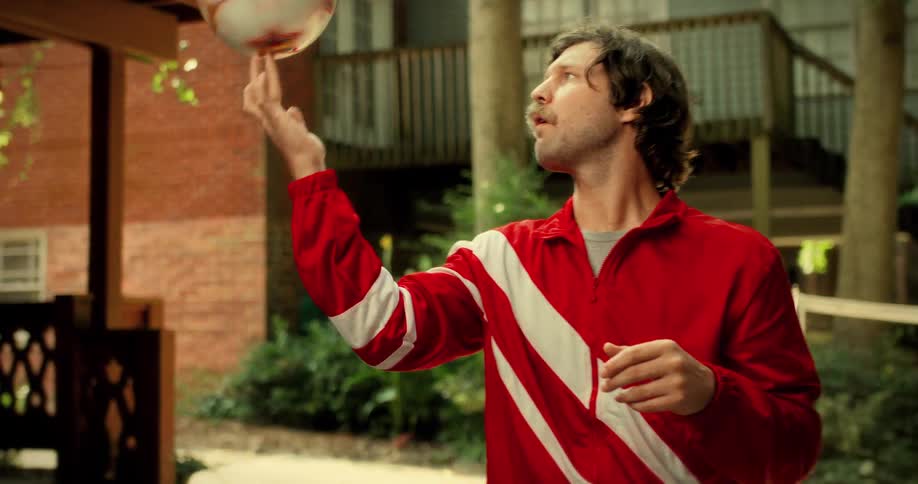 A mustached man in a retro red tracksuit spins a soccer ball on his finger in slow motion in a backyard setting