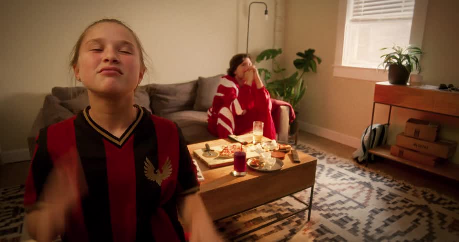 An intensely focused young girl in a soccer jersey watches a tense game at home while her dad watches from the couch
